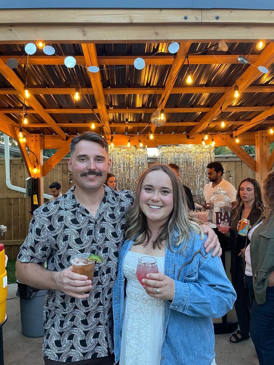 Couple holding cocktails under string lights at a backyard bar
